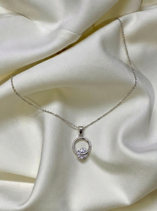 Zoe Solitaire Pendant Necklace featuring a delicate silver chain and a circular halo pendant setting with a central round brilliant solitaire stone on draped white satin