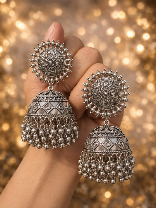 Isitaara detailed Maharani oxidized silver Jhumka earrings featuring large, circular textured mandala studs and hanging metallic bead bells held in a hand against a warm, golden bokeh background.

