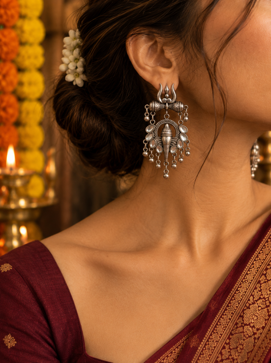 A close-up lifestyle shot of a woman wearing an ornate silver-toned traditional Indian jhumka earring. The earring features intricate filigree work, hanging silver beads, and a central bell-shaped charm.